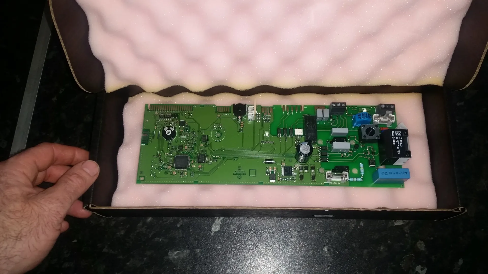 A green circuit board with various electronic components, possibly a boiler PCB, packaged in a box with pink foam.